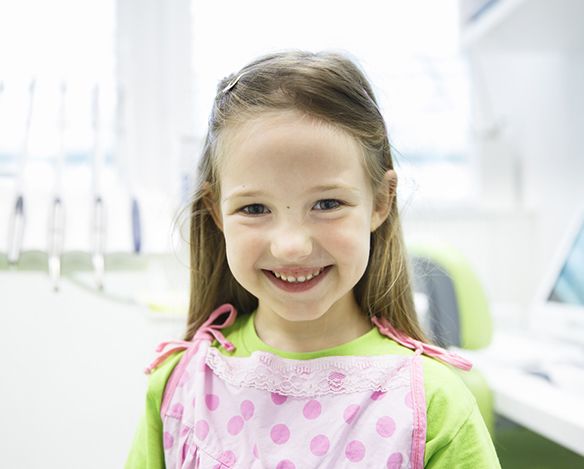 A little girl wearing a pink apron and green shirt smiles after receiving tooth-colored fillings in Coppell