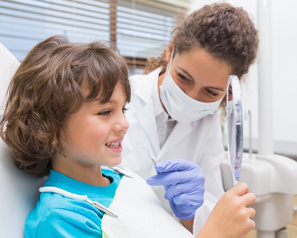 A dentist smiles behind a mask while a little boy holds a handheld mirror and looks at his teeth