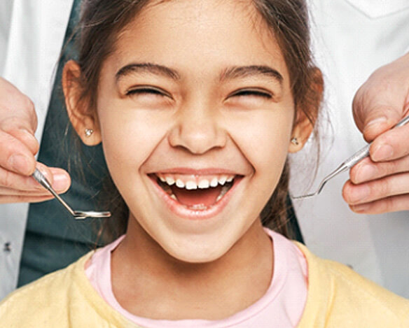 A young girl smiles wide while a dentist stands behind her with dental instruments in preparation for checking her smile