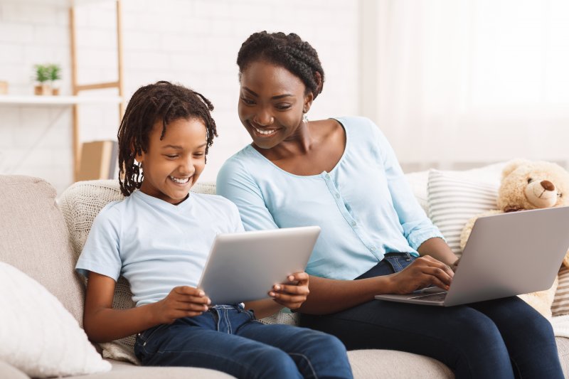 a mother and her child sitting on the couch on their tablet and computer smiling while at home