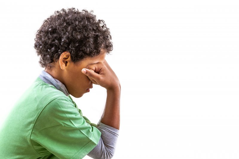 a little boy wearing a green t-shirt sitting with his fisted hands resting on his eyes as if he is stressed